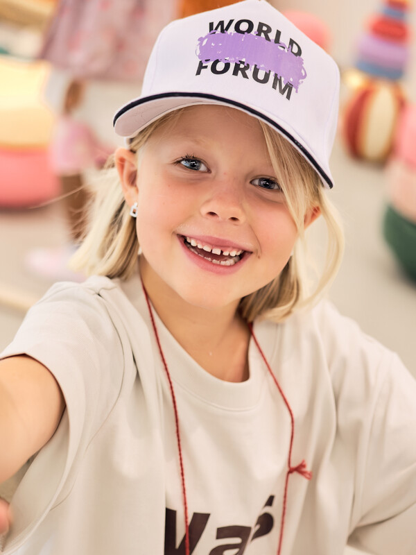 A young participant in a World Child Forum cap smiles and reaches toward the camera
